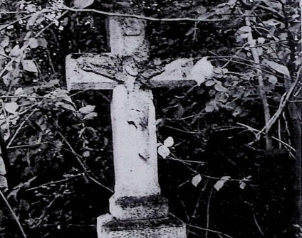 Nagrobek z krzyżem na dwustopniowym cokole otoczony roślinnością. Krzyż ma ścięte krawędzie i relief Chrystusa na przecięciu ramion. Powyżej titulus.