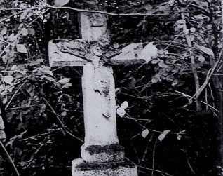 Nagrobek z krzyżem na dwustopniowym cokole otoczony roślinnością. Krzyż ma ścięte krawędzie i relief Chrystusa na przecięciu ramion. Powyżej titulus.