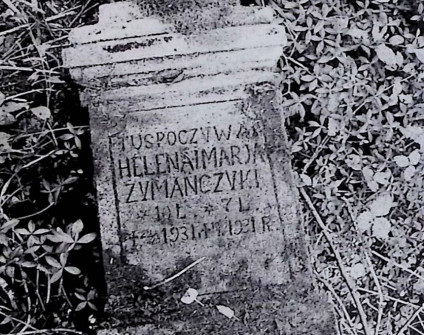 Nagrobek Heleny i Marii Żymańczyk leżący na ziemi. Napis: 'Tu spoczywają Helena i Maria Żymańczyk'. Otoczony trawą i roślinami.