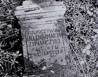 Nagrobek Heleny i Marii Żymańczyk leżący na ziemi. Napis: 'Tu spoczywają Helena i Maria Żymańczyk'. Otoczony trawą i roślinami.