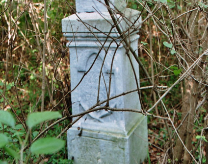 Kamienny cokół nagrobka z wyrytymi inskrypcjami, częściowo zasłonięty gałęziami. Cokół ma profilowane górne krawędzie i łukowatą płycinę z przodu.