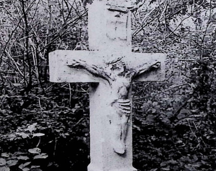 Kamienny krzyż z reliefem Chrystusa na przecięciu ramion, otoczony gęstą roślinnością. Powyżej postaci widoczny titulus.