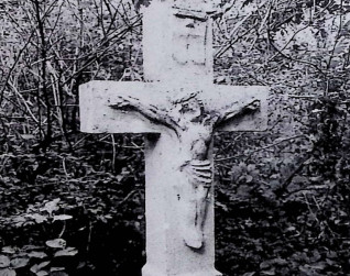 Kamienny krzyż z reliefem Chrystusa na przecięciu ramion, otoczony gęstą roślinnością. Powyżej postaci widoczny titulus.