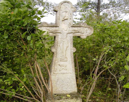 Kamienny krzyż na cokole z ramionami zakończonymi trójliściem, z płaskorzeźbą Chrystusa, titulus i czaszką z piszczelami. Otoczony zielenią.