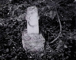 Stary nagrobek z utrąconym krzyżem na cokole, otoczony roślinnością. Krzyż ma owalną niszę z płaskorzeźbionym czteroliściem. Cokół ma profilowane krawędzie i wyrytą ramę.