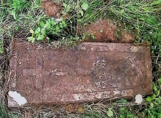 Betonowy nagrobek Michała Przyjemskiego z tablicą o ćwierćkoliście ściętych narożach i krzyżem, z wyrytą inskrypcją, otoczony trawą.