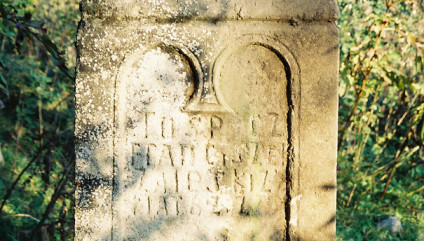 Kamienny cokół o profilowanych krawędziach z płyciną zamkniętą łukiem i inskrypcją. Krzyż niezachowany. Powierzchnia nosi ślady erozji.