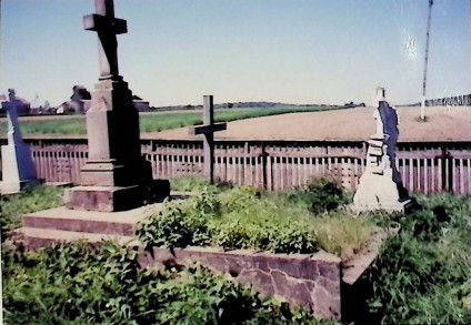 Nagrobek z krzyżem na cokole i dwustopniowej podstawie, otoczony trawą. Na krzyżu relief Chrystusa i napis 'I.N.R.I.' W tle drewniany płot i otwarte pola.