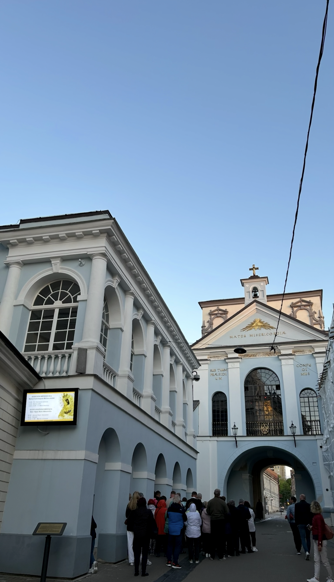The Gate of Dawn in Vilnius with its classical facade and chapel. A group of people stands beneath the archway. The facade features inscriptions in Latin.