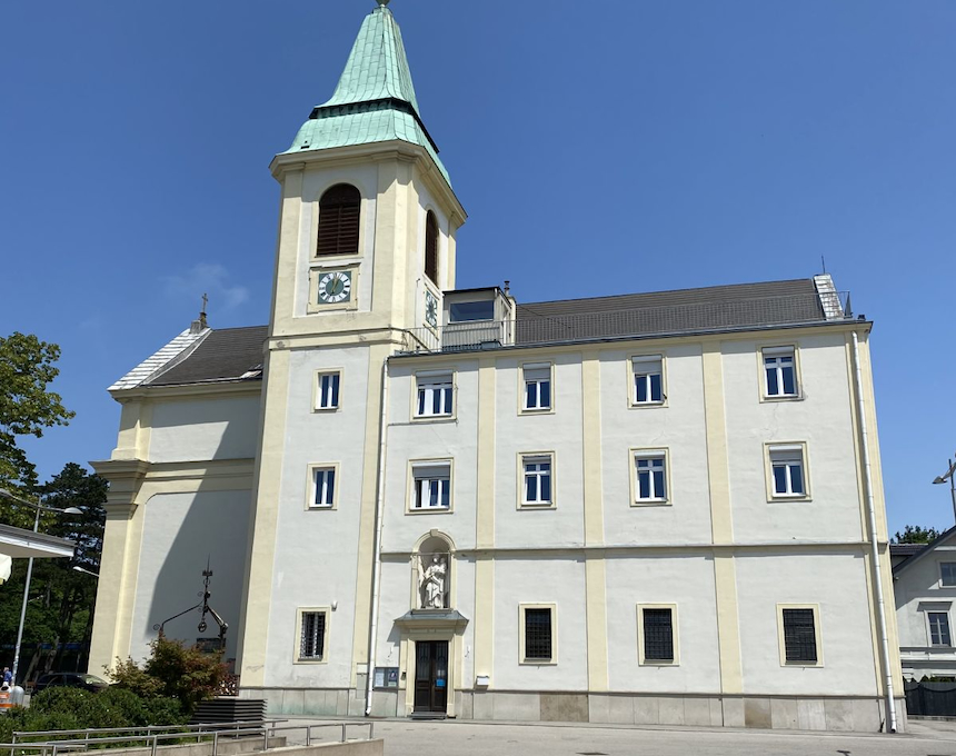 Façade de l'église située sur la colline du Kahlenberg à Vienne, avec une tour d'horloge au toit vert et plusieurs fenêtres. Le bâtiment est de couleur claire et une statue se trouve au-dessus de l'entrée.