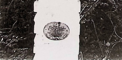 Grób oznaczony krzyżem stylizowanym na pień drzewa na cokole z naprzemiennie ułożonych drewnianych belek. Owalna metalowa tablica z wypukłą inskrypcją i skrzyżowanymi liśćmi palmowymi u podstawy.