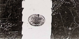 Grób oznaczony krzyżem stylizowanym na pień drzewa na cokole z naprzemiennie ułożonych drewnianych belek. Owalna metalowa tablica z wypukłą inskrypcją i skrzyżowanymi liśćmi palmowymi u podstawy.