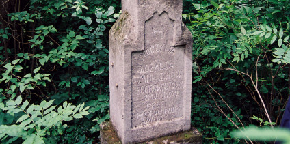 Nagrobek Rozalii Fedorowicz z krzyżem na cokole i dwustopniowej podstawie, z płyciną z falistym górnym brzegiem i inskrypcją, otoczony zielenią.