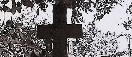 Grób z krzyżem na cokole, ozdobionym guzkami i rozetami. Na cokole wyryte serce z krzyżykiem i oszklona tablica z inskrypcją.