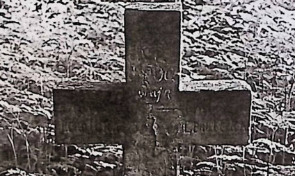 Grób oznaczony prostym krzyżem z wyrytą inskrypcją, otoczony trawą.