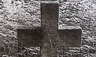 Grób oznaczony prostym krzyżem z wyrytą inskrypcją, otoczony trawą.