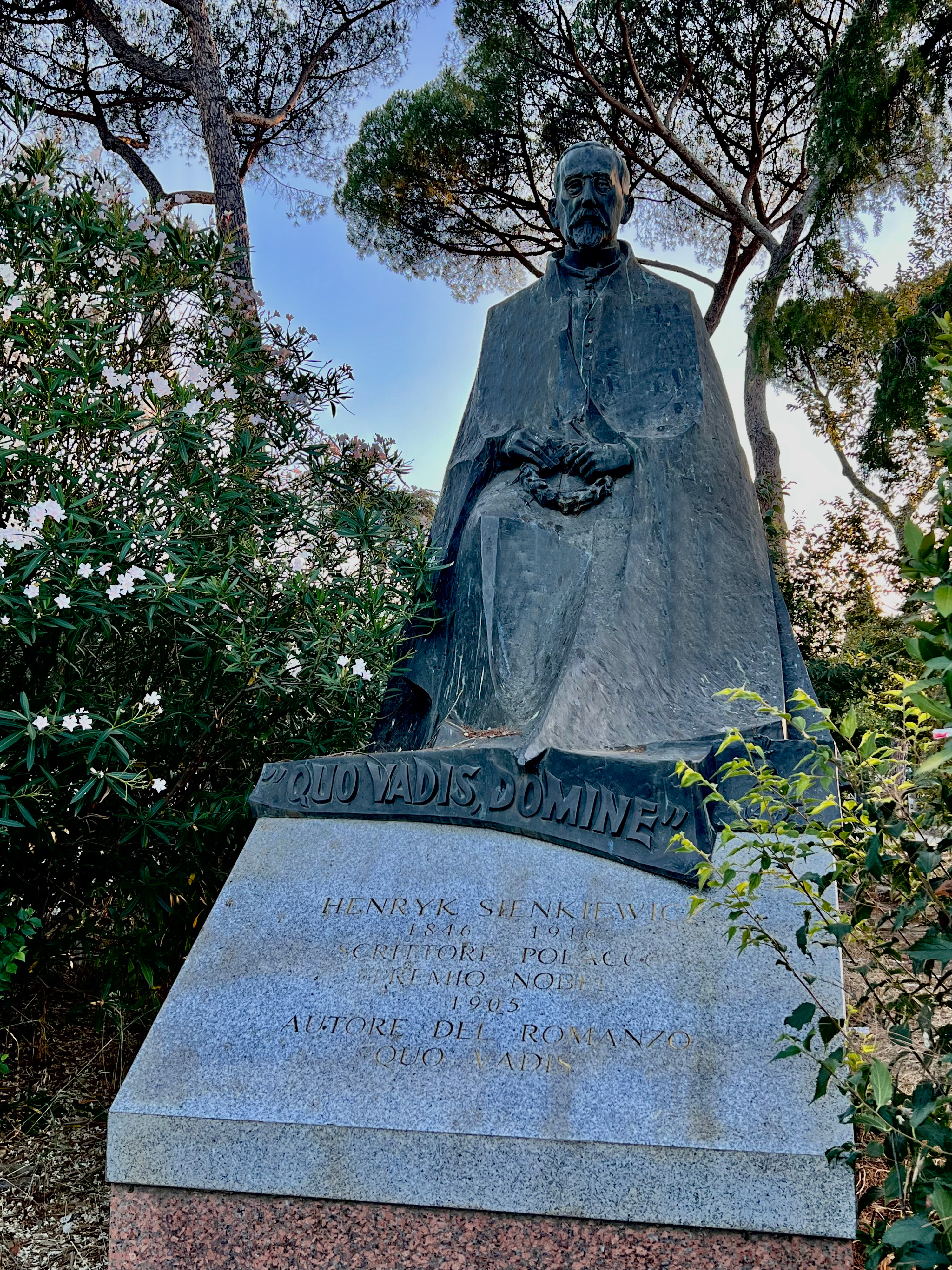 Statue of Henryk Sienkiewicz holding a laurel wreath, with the inscription 'Quo Vadis Domine' at the base. Surrounded by trees and flowers in Villa Borghese Gardens.