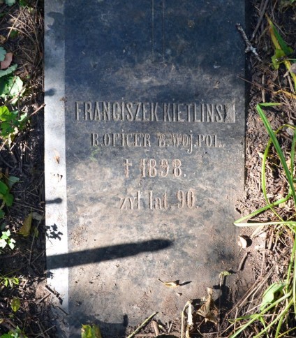 Polerowany nagrobek Franciszka Kietlińskiego o ściętych krawędziach, z napisem i symbolem krzyża. Otoczony trawą i ziemią.