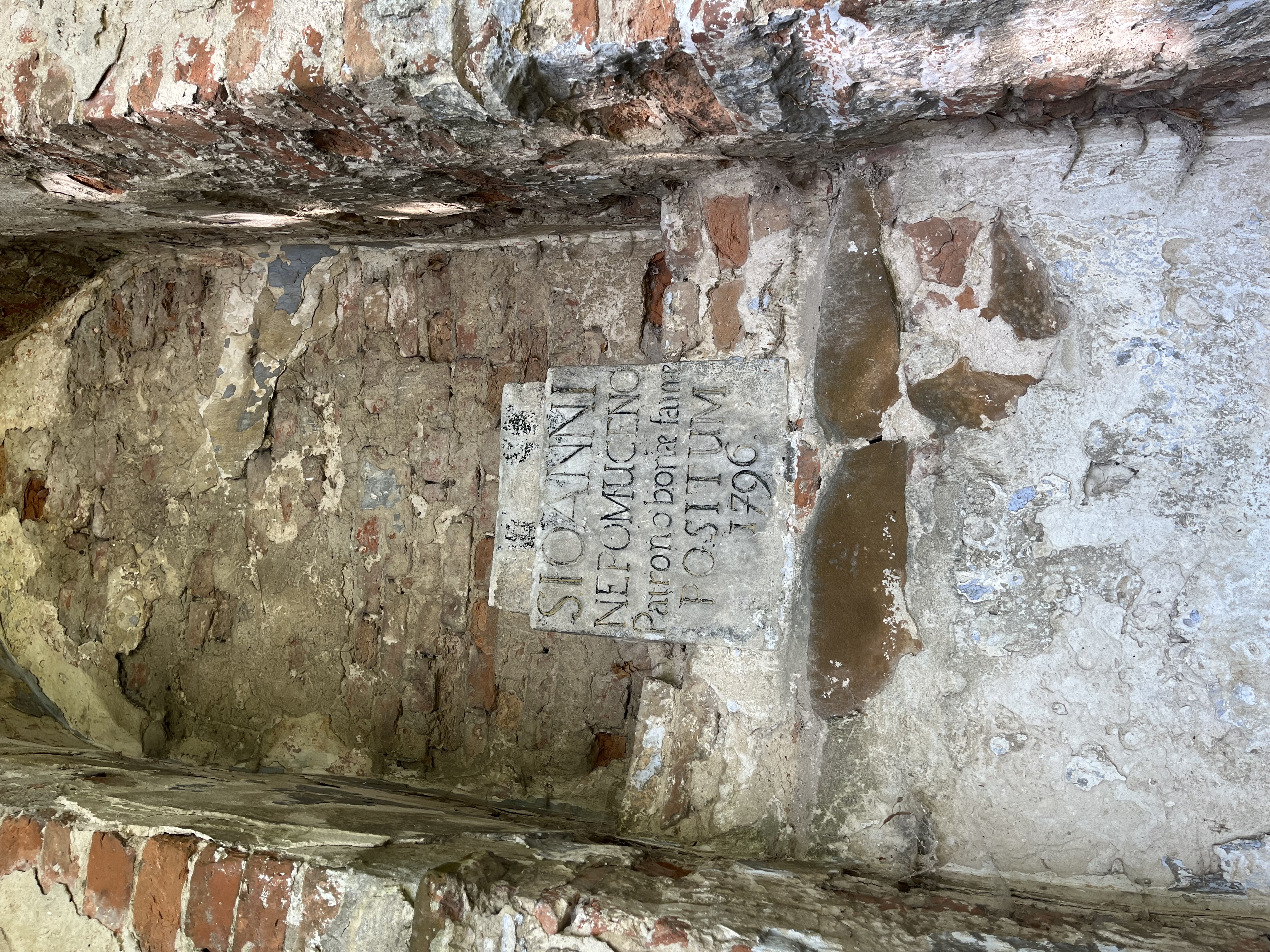 Old brick wall with a weathered plaque inscribed with 'S. IOANNI NEPOMUCENO Patrono bonae famae POSITUM 1796' at the cemetery in Kościejów.