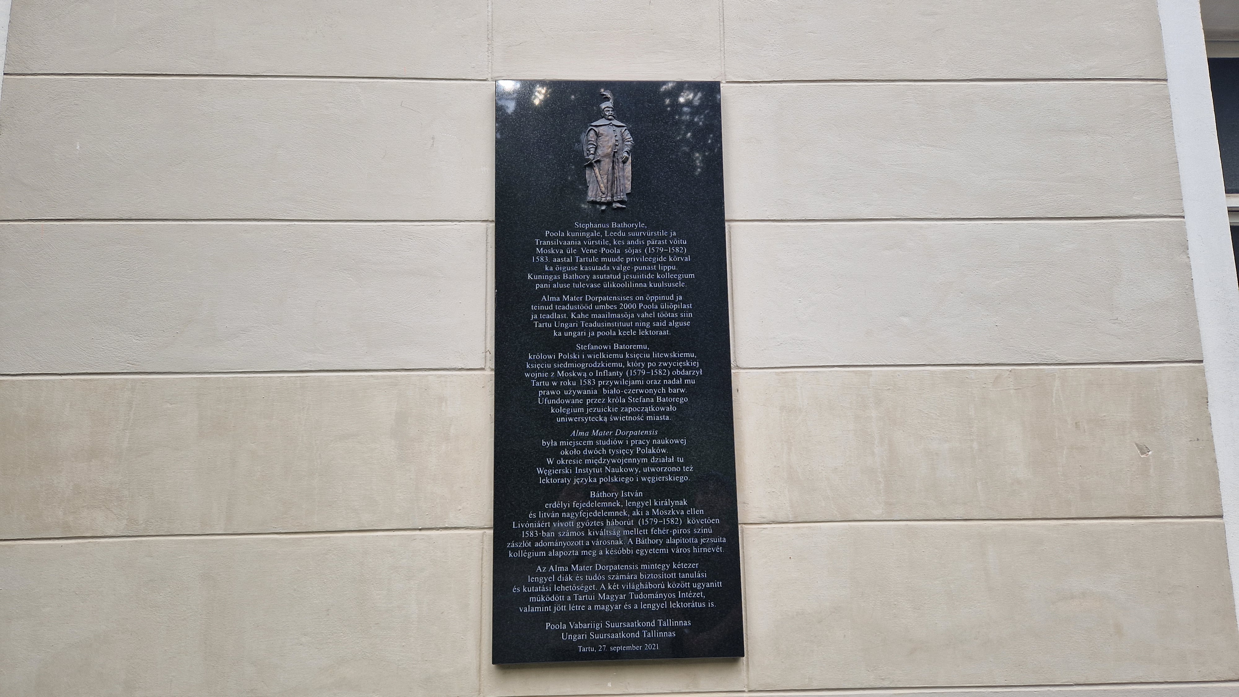 Пам'ятна дошка на бежевій стіні з рельєфом чоловіка в традиційному вбранні. Текст естонською та польською мовами вшановує короля Стефана Баторія за заснування єзуїтського коледжу в Тарту в 1583 році.