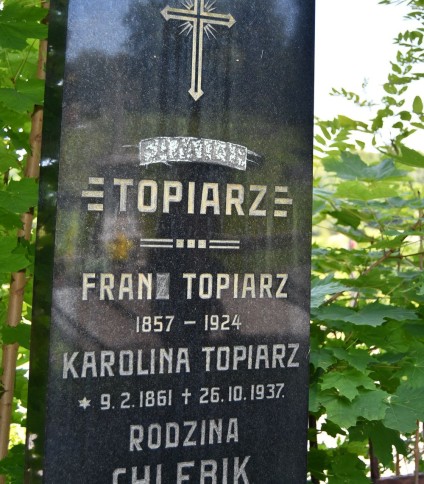 Nagrobek rodziny Topiarzów z krzyżem i inskrypcjami dla Frana Topiarza (1857-1924) i Karoliny Topiarz (1861-1937). Otoczony zielenią.