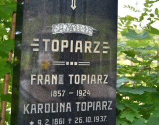 Nagrobek rodziny Topiarzów z krzyżem i inskrypcjami dla Frana Topiarza (1857-1924) i Karoliny Topiarz (1861-1937). Otoczony zielenią.
