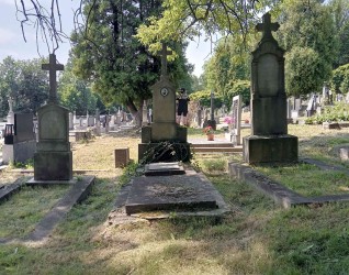 Grób oznaczony krzyżem na dwustopniowym cokole i podstawie, z tablicą inskrypcyjną i pozostałościami krucyfiksu. Otoczony drzewami i innymi grobami na cmentarzu.