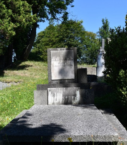 Nagrobek rodziny Kamockich z pionową tablicą na cokole, z wyrytymi inskrypcjami. Całość ustawiona na trójstopniowej podstawie, otoczona trawą i drzewami.