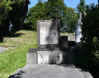 Nagrobek rodziny Kamockich z pionową tablicą na cokole, z wyrytymi inskrypcjami. Całość ustawiona na trójstopniowej podstawie, otoczona trawą i drzewami.