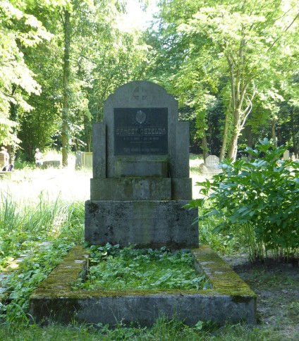 Nagrobek Ernesta Oszelja z stelą na cokole, z okrągłym medalionem z krzyżem i tablicą z inskrypcją. Pole grobowe ujęte opaską, otoczone zielenią.