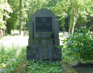 Nagrobek Ernesta Oszelja z stelą na cokole, z okrągłym medalionem z krzyżem i tablicą z inskrypcją. Pole grobowe ujęte opaską, otoczone zielenią.