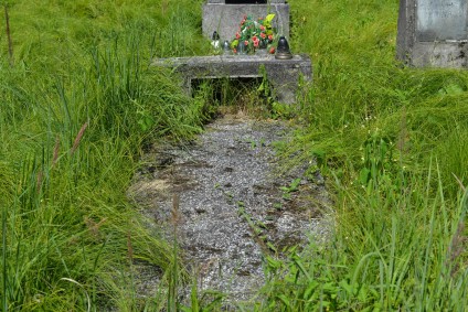 Miejsce pochówku ze stelą na cokole i podstawie, otoczone trawą. Stela poprzedzona płytą nakrywającą piwnicę grobową. W przestrzeni widoczne fragmenty szkła.