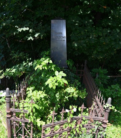 Grób Joanny Hausslerowej z wypolerowanym obeliskiem z inskrypcją z przodu. Grób otoczony metalowym ogrodzeniem z dekoracjami geometrycznymi i furtką. Otoczenie porośnięte roślinnością.