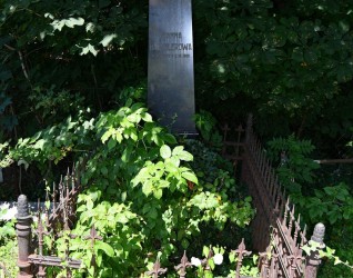 Grób Joanny Hausslerowej z wypolerowanym obeliskiem z inskrypcją z przodu. Grób otoczony metalowym ogrodzeniem z dekoracjami geometrycznymi i furtką. Otoczenie porośnięte roślinnością.