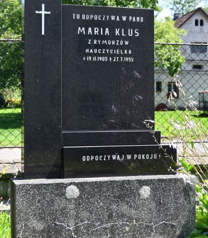 Nagrobek Marii Klus z czarną stelą z monzonitu z inskrypcjami i krzyżem. Podstawa z lastryko. W tle zieleń i dom.