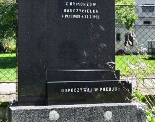 Nagrobek Marii Klus z czarną stelą z monzonitu z inskrypcjami i krzyżem. Podstawa z lastryko. W tle zieleń i dom.