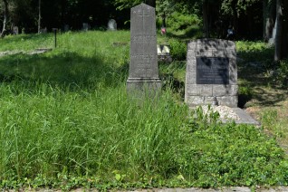Grób oznaczony marmurowym obeliskiem na betonowym cokole z krzyżem i inskrypcją dla Jerzego Kobieli, Anny Podlowskiej i Zuzanny Kocharczyk, otoczony trawą.