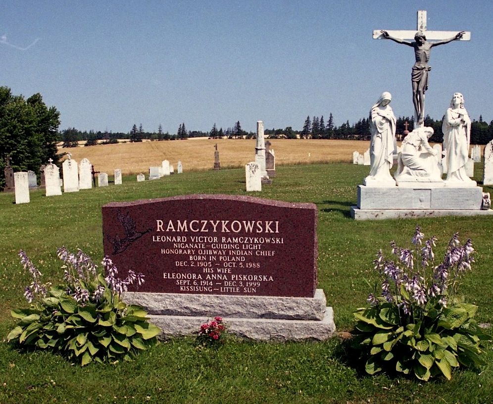Nagrobek Leonarda Victora Ramczykowskiego i Eleonory Anny Piskorskiej na cmentarzu. Kamień otoczony roślinami. W tle widoczna biała figura sceny ukrzyżowania.