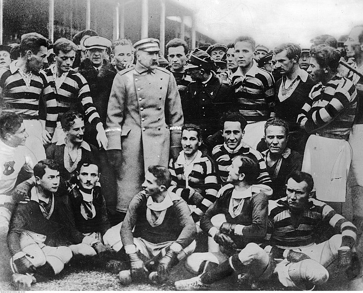 Historyczna fotografia drużyny piłkarskiej Lwowskiego Klubu Sportowego 'Pogoń', z zawodnikami w pasiastych koszulkach i spodenkach, zgromadzonymi wokół mężczyzny w mundurze wojskowym. Grupa znajduje się na zewnątrz, w tle tłum.