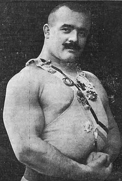 Stanisław Zbyszko Cyganiewicz, muskularny mężczyzna z wąsem, pozuje w stroju zapaśniczym ozdobionym medalami i wstążkami.