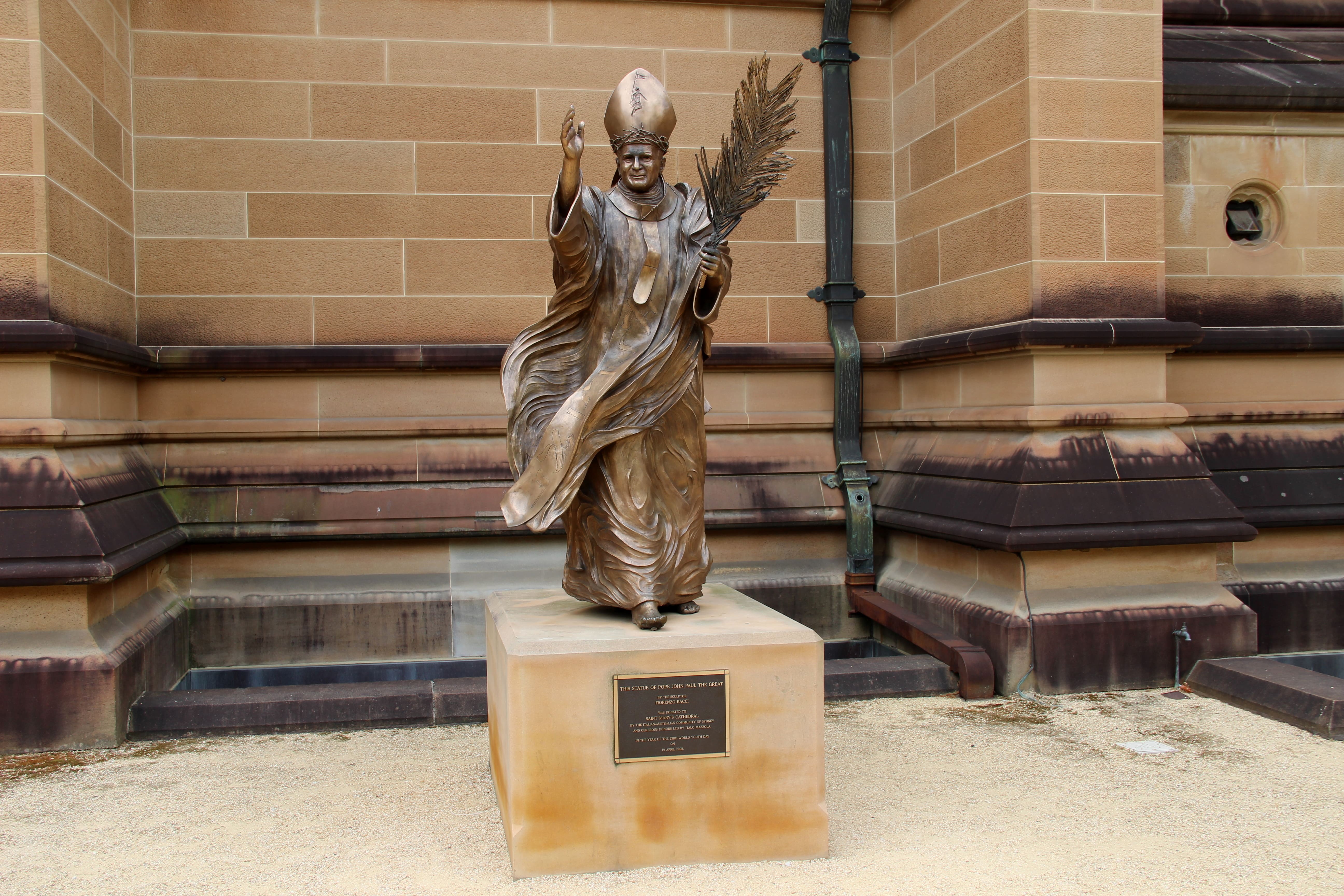 Brązowa statua Jana Pawła II trzymającego gałązkę palmową, z jedną ręką uniesioną w geście błogosławieństwa. Statua stoi na kamiennym cokole z inskrypcją, przy ceglanej ścianie.