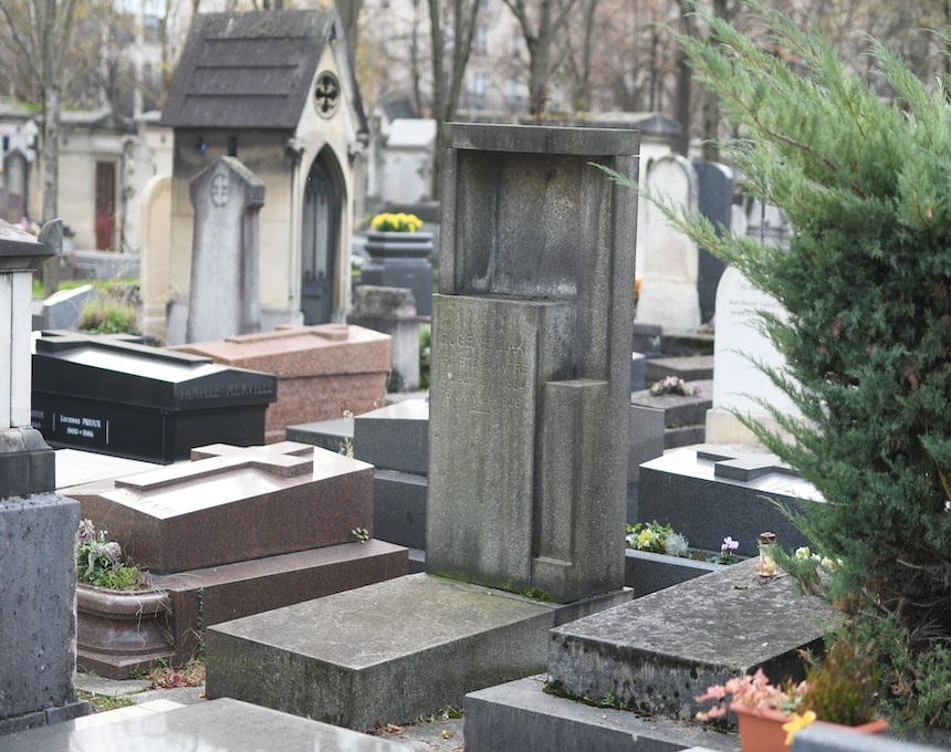 Grób Eugeniusza Zaka na cmentarzu Montparnasse w Paryżu. Modernistyczny nagrobek z szarego kamienia z prostokątną wnęką i inskrypcją. Widoczne otaczające groby i zieleń.