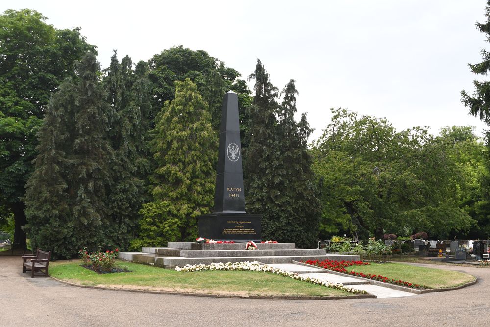 Czarny obelisk z napisem 'Katyń 1940' stoi otoczony drzewami i kwiatami. Jest częścią cmentarza, z ławką i kwiatowymi aranżacjami w pobliżu.