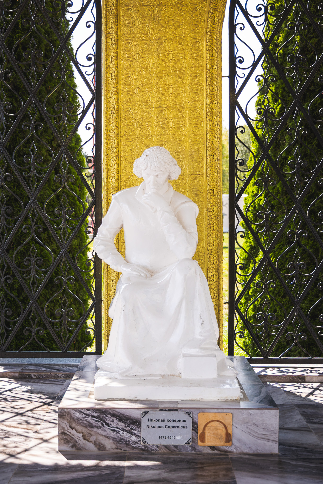 Biała statua Mikołaja Kopernika siedzącego w zamyślonej pozie, z ręką na brodzie, na tle złotej ornamentyki. Na podstawie tabliczka z jego imieniem w cyrylicy i łacinie oraz daty urodzenia i śmierci.