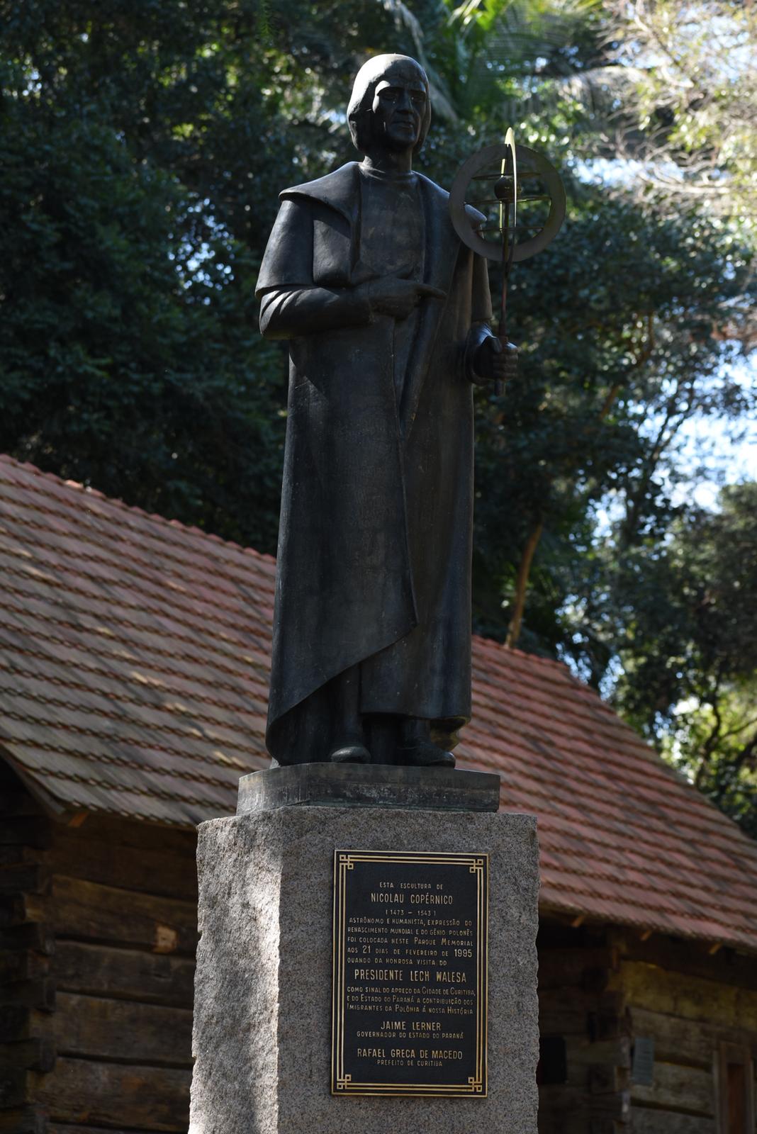Bronze statue of Nicolaus Copernicus on a stone pedestal with a plaque in Curitiba, Brazil. The statue depicts Copernicus holding an astrolabe, set against a backdrop of trees and a wooden building.