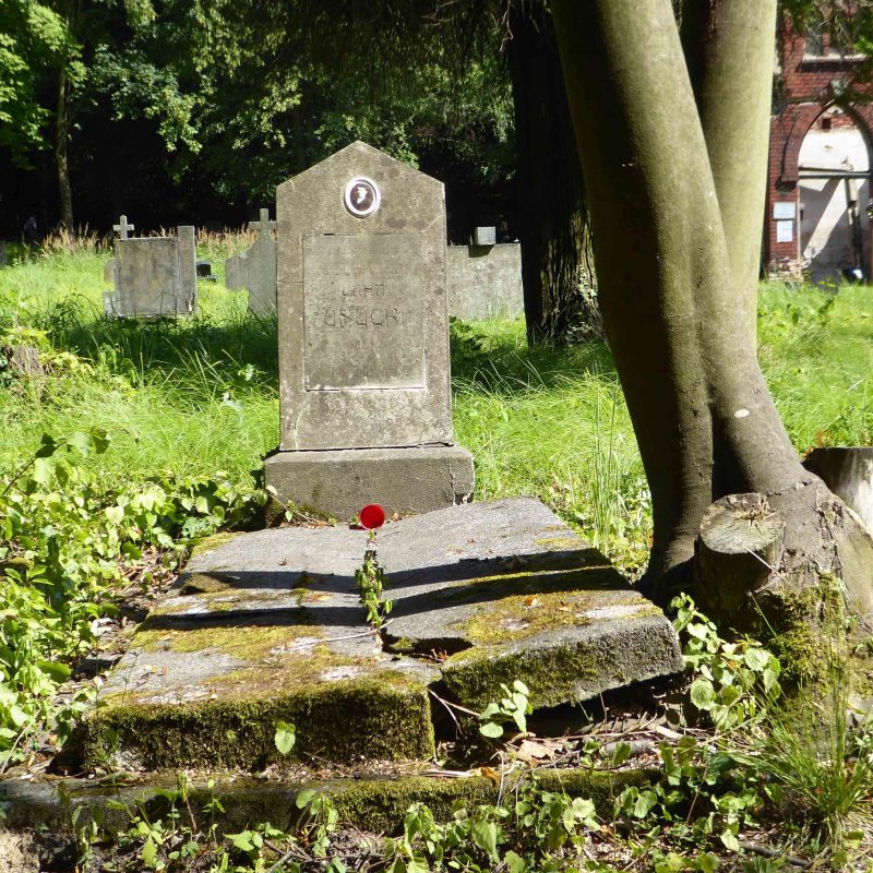 Nagrobek Jana Unuckiego z dwuspadową stelą na cokole, z owalnym medalionem fotograficznym i tablicą z inskrypcją. Grób otoczony zielenią i drzewami.