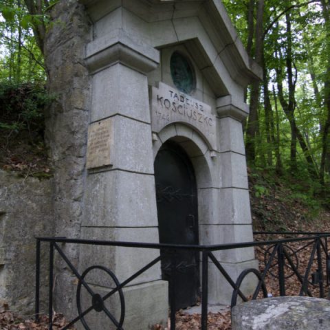 Кам'яна структура, схожа на каплицю, з арочними чорними дверима, оточена металевою огорожею. Напис над дверима: 'Тадеуш Костюшко 1746-1817'. Розташована в лісистій місцевості.