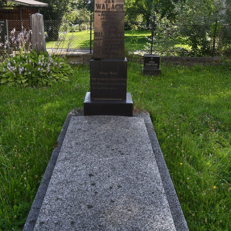 Grób rodziny Walach z wypolerowanym obeliskiem z inskrypcjami i krzyżem. Pole grobowe ujęte opaską i nakryte płytą. Widoczna otaczająca zieleń i drzewa.