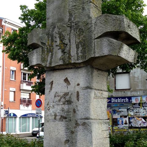 Monument en pierre à Lille avec des blocs verticaux et horizontaux croisés, portant une inscription en français et en polonais. Entouré de verdure et de bâtiments urbains.