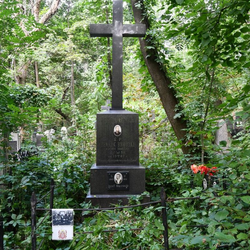 Grave of Henryk Brodzki and Józef Buczyński marked by a cross on a three-tiered pedestal with engraved inscriptions and ceramic medallions featuring male portraits. Surrounded by a metal fence with a family photo and coat of arms.